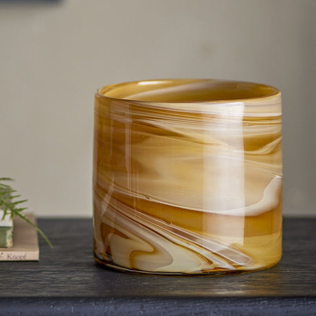 Sylas Glass Tealight Holder in amber, a round cylindrical glass votive with marbled swirl patterns, shown on a dark shelf against a neutral background.
