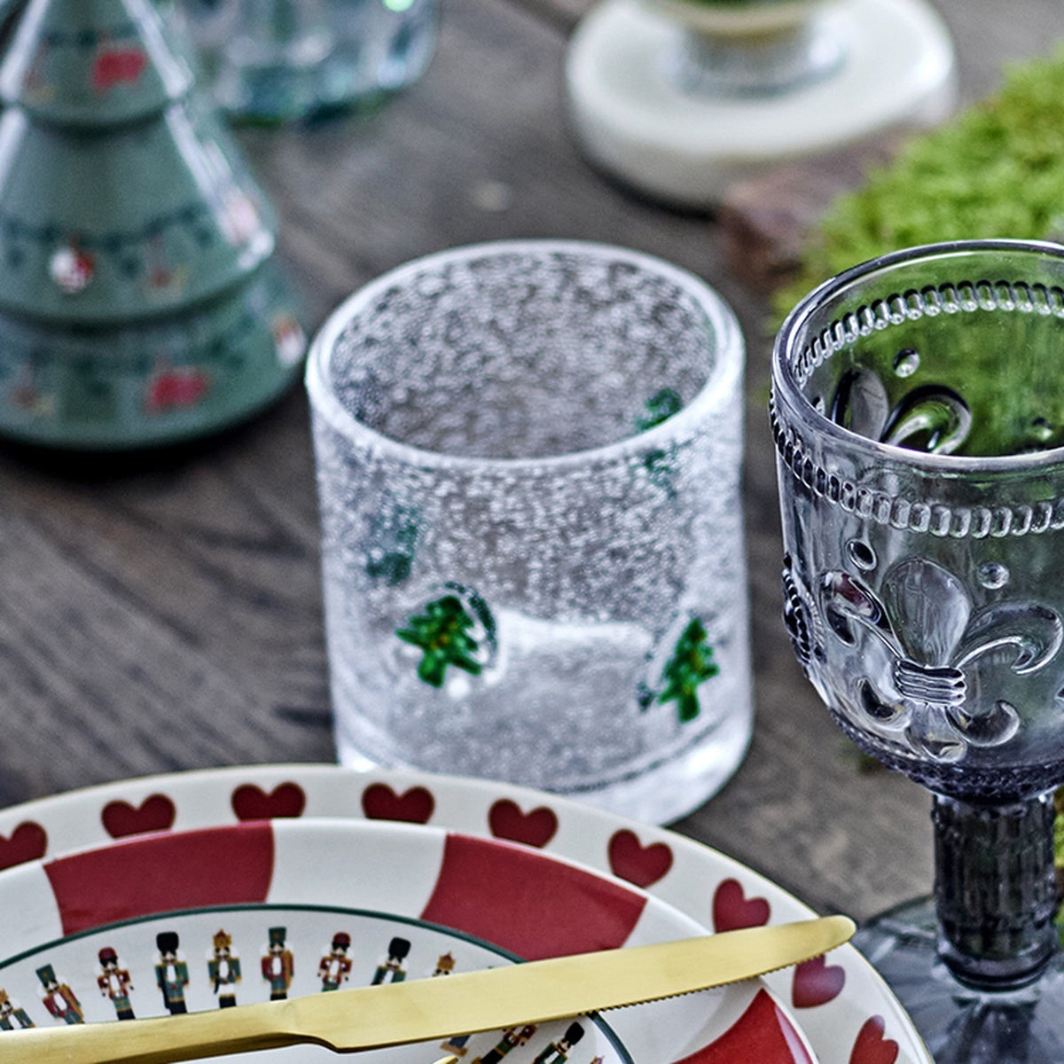 Debra Christmas Tree Tumbler in clear bubbled glass with hand-painted green Christmas trees, styled on a wooden table beside a grey patterned stem glass and festive plates.