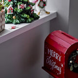 Carlo Christmas Decoration in red MDF, a postbox-style ornament with slatted curved roof, letter slot, "Merry Christmas" script, and a small wreath, displayed near a festive mantel.
