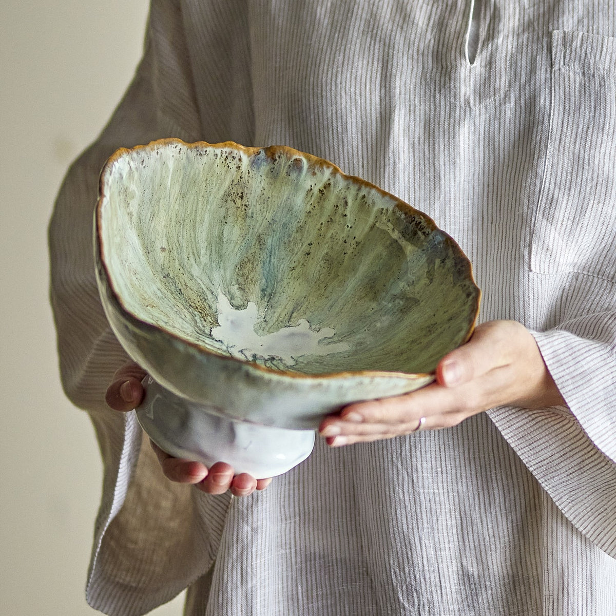 Joby Stoneware Bowl 24cm in Green, a footed ceramic bowl with an irregular wavy rim and streaked green glaze, held in two hands against a neutral background.