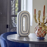 Mae Striped Ceramic Vase 32cm in white, an upright oval loop shape with navy outline stripes, displayed on a table beside a gold candelabra.