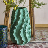 Natika Wavy Terracotta Vase 37cm in green with ruffled, wavy ridges, shown holding foliage in a room with a mosaic glass votive and wooden easel nearby.