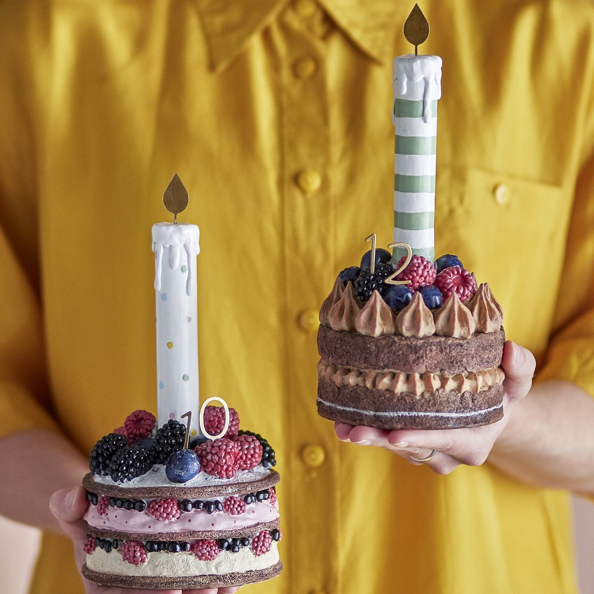 Hooray Resin Birthday Decorations Set of 2 held by hand, berry-topped layer cake designs with striped and dotted candles, gold flames, numbers 10 and 12.