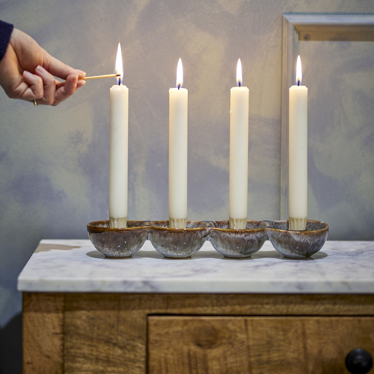 Kalia Stoneware Advent Candle Holder in brown reactive glaze, four joined round cups holding lit ivory taper candles, shown on a marble-top wooden table as a hand lights one with a match.