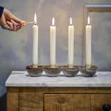 Kalia Stoneware Advent Candle Holder in brown reactive glaze, four joined round cups holding lit ivory taper candles, shown on a marble-top wooden table as a hand lights one with a match.