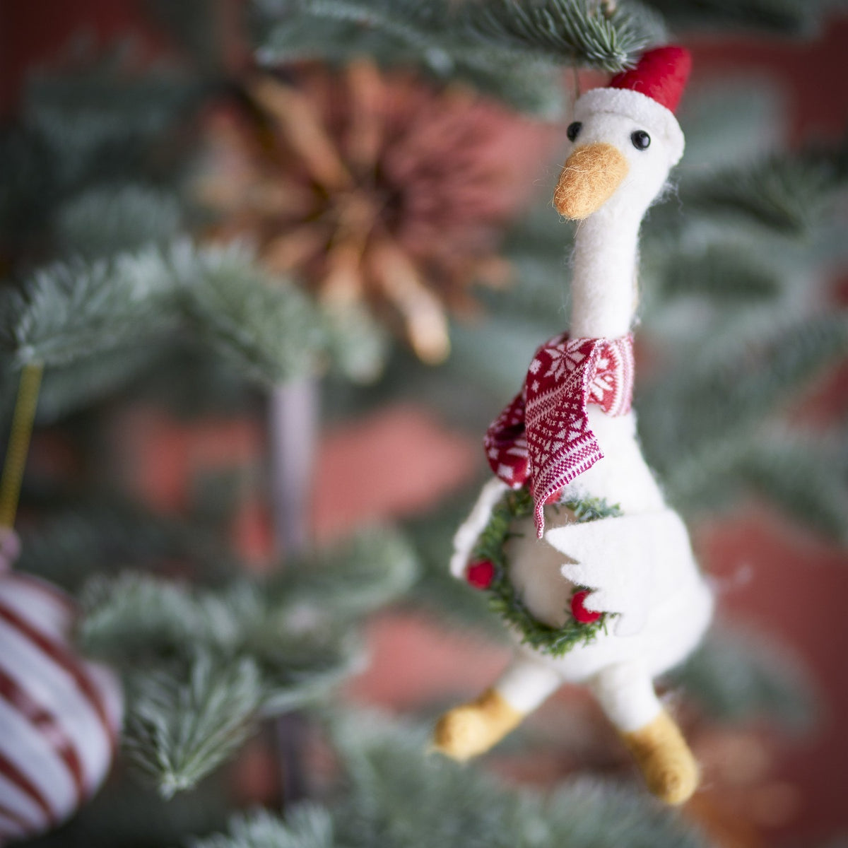Arlo Wool Christmas Tree Decoration in white felt, a goose figure wearing a red Santa hat and snowflake scarf, holding a berry wreath, hanging among pine branches.