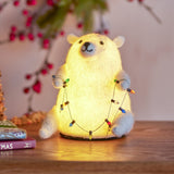 White felted wool Wally bear Christmas decoration with built-in light, holding a bead and bulb garland, displayed on wood with berry sprigs behind it.