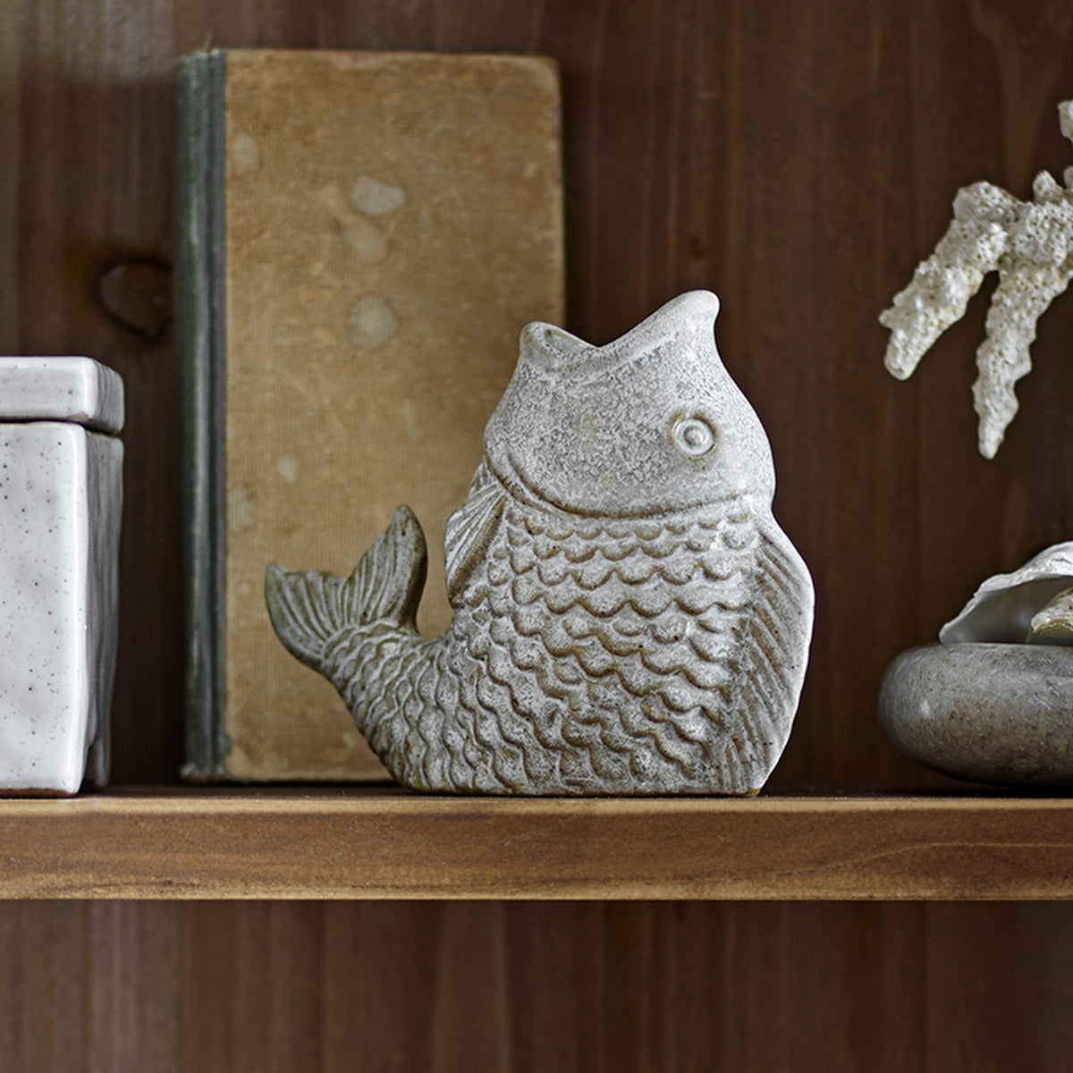 Hiro Fish Stoneware Candle Holder in grey, shown upright with textured scales and curled tail, displayed on a wooden shelf beside books and decor.
