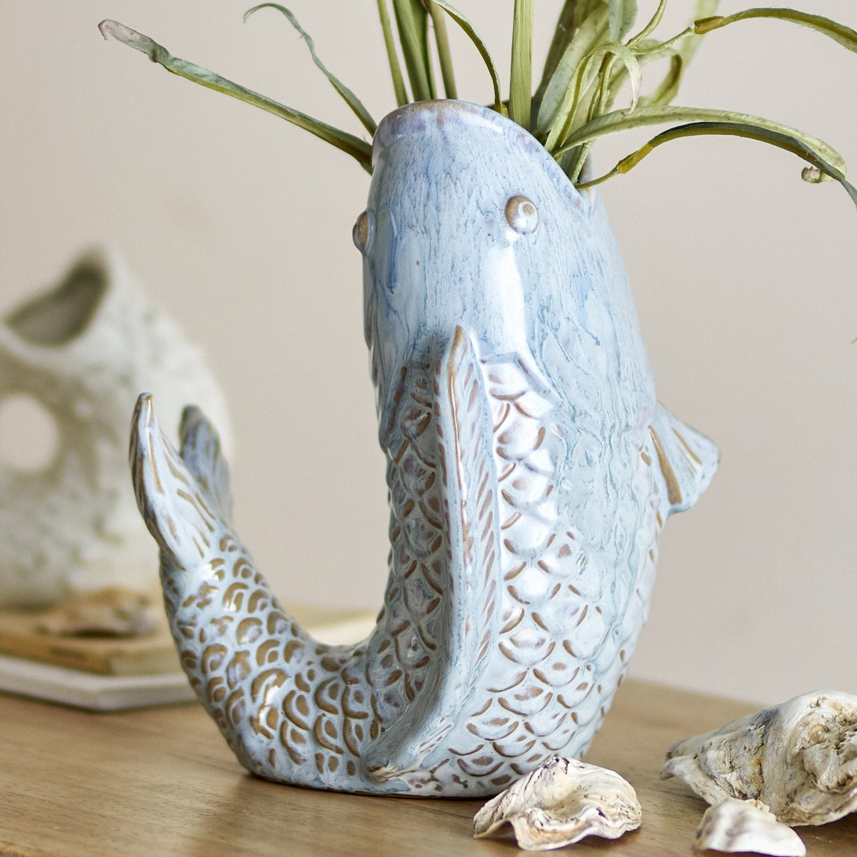 Hiro Fish Stoneware Vase 21cm in blue, shaped like a leaping fish with scaled body, curled tail and open mouth forming the neck, holding trailing greenery on a wooden surface.
