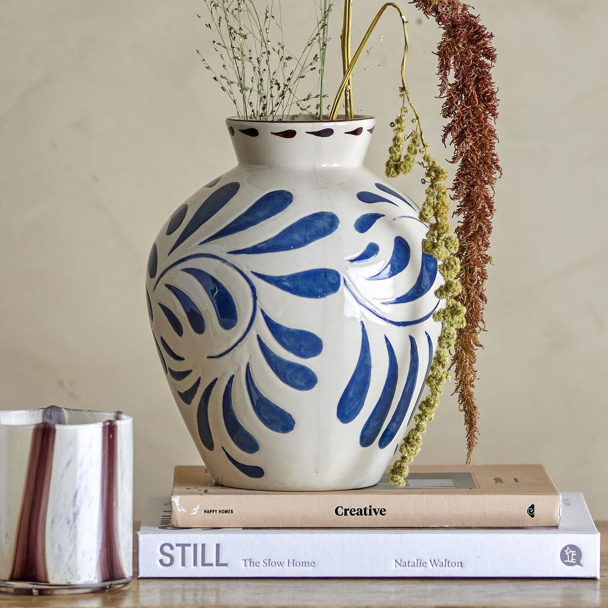 Heikki Stoneware Vase 24cm in cream with bold blue hand-painted leaf motifs, shown with dried foliage atop stacked books in a living room setting.