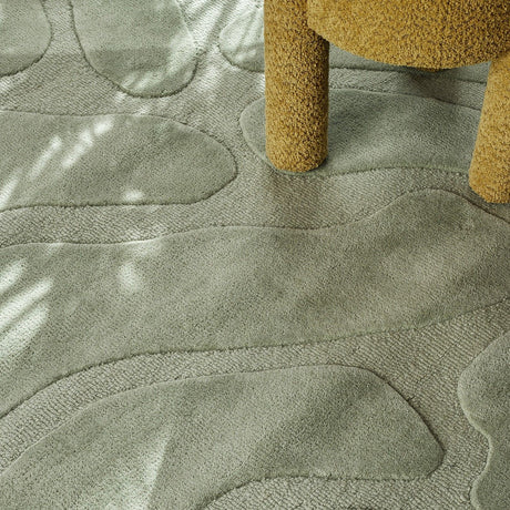 Darlington Wool Rug in green, 200x130cm, showing tufted pebble-like shapes in raised pile against lower, textured channels, with a boucle chair leg resting on the surface.
