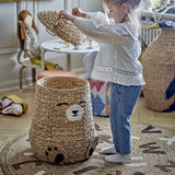 Timone Bear water hyacinth storage basket with lid in natural finish, shaped like a sitting bear with stitched face and paw details, shown in a child's playroom as a girl lifts the lid.