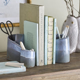 Meliad Stoneware Bookends Set of 2 in blue, curled hollow ceramic forms with glossy light blue rims fading to speckled dark blue, shown holding books, scissors, and pencils on a wooden shelf.