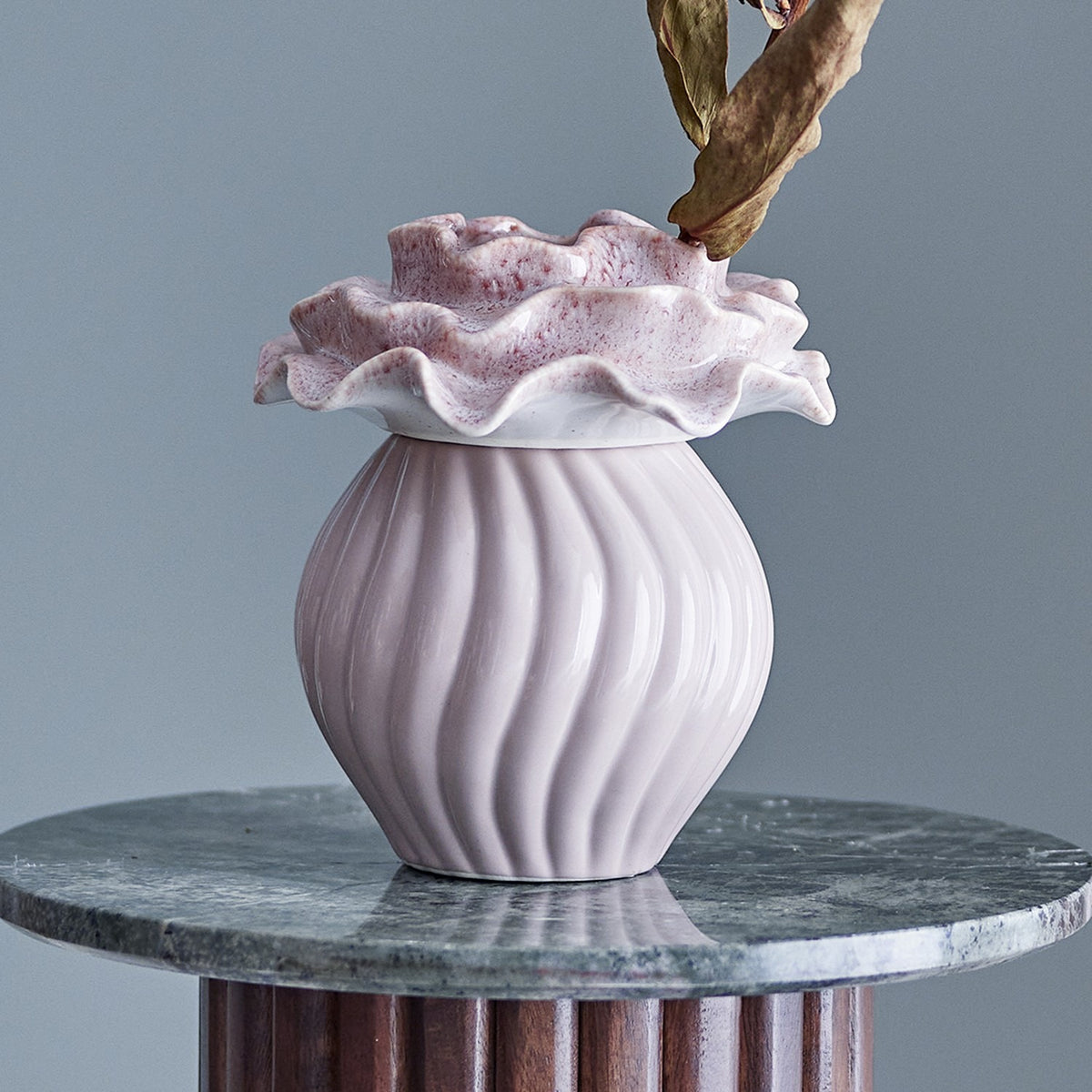Petalia Stoneware Vase 17cm in pink, featuring a fluted, ribbed round body in glossy pink ceramic topped with a ruffled, speckled petal-like collar, shown on a marble surface with a dried leaf stem.
