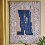 Atle Wool Wall Decor in multi tones, a tufted wool hanging with a navy figure bent forward in long hair, framed by a grey leafy sprig border, shown mounted on a wall.