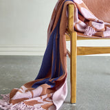 Atle Leaf Cotton Throw in brown with lilac leaf print and navy and tan stripes, cotton weave with tasselled fringe, draped over a wooden chair with tan leather seat.