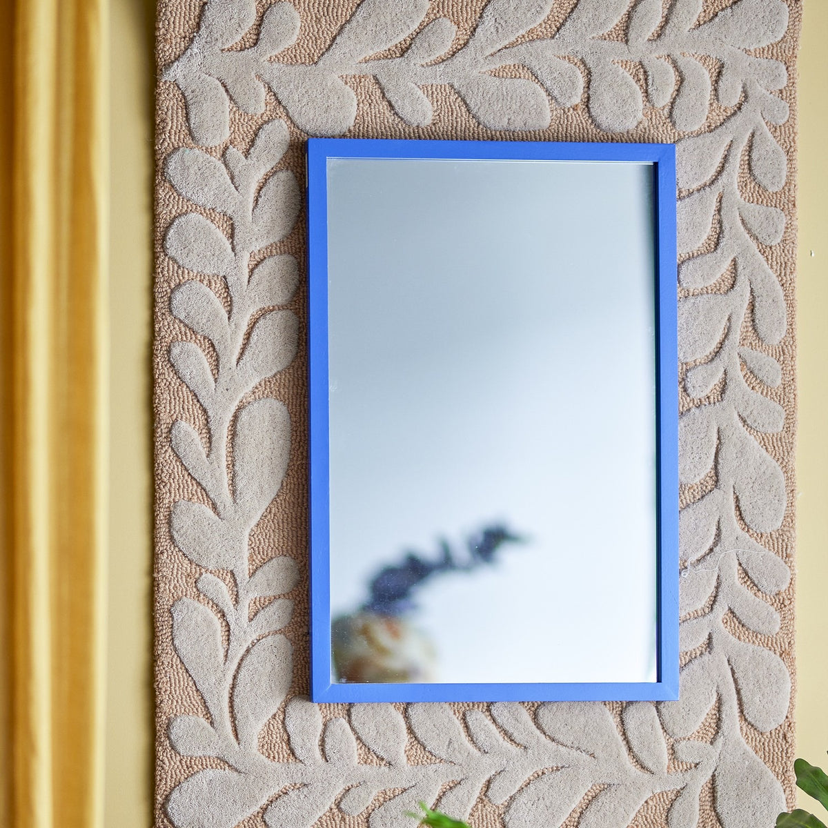 Atle Wall Mirror 40x60cm in Blue, a rectangular MDF-framed mirror mounted on a textured beige leaf-patterned wall panel beside a gold curtain.
