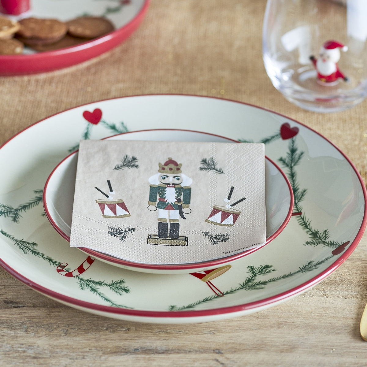 Beige paper napkin from the Jolly Christmas pack of 20, printed with a nutcracker soldier and drums design, folded on a festive plate setting.