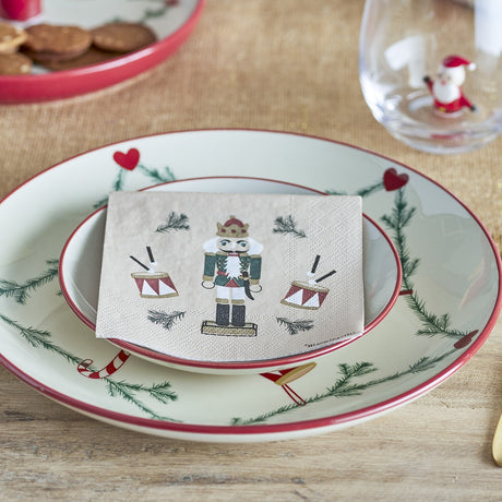 Beige paper napkin from the Jolly Christmas pack of 20, printed with a nutcracker soldier and drums design, folded on a festive plate setting.