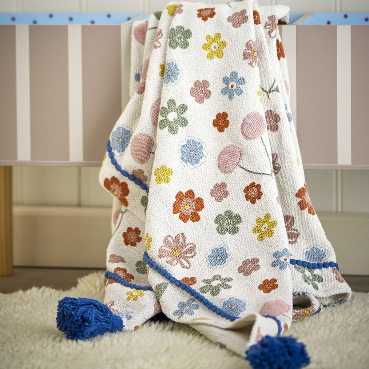 Cherry Floral Cotton Throw in multicoloured cotton blend, draped over a striped cot with blue tasselled corners, displayed on a sheepskin rug in a nursery setting.