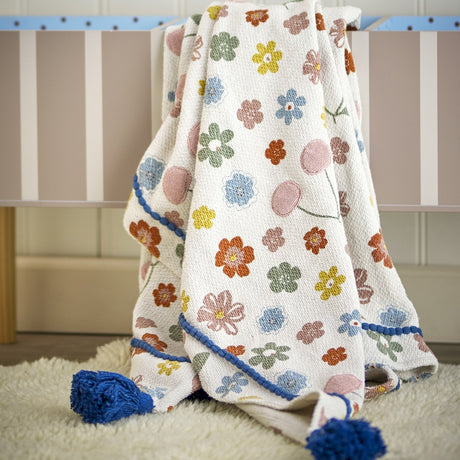Cherry Floral Cotton Throw in multicoloured cotton blend, draped over a striped cot with blue tasselled corners, displayed on a sheepskin rug in a nursery setting.