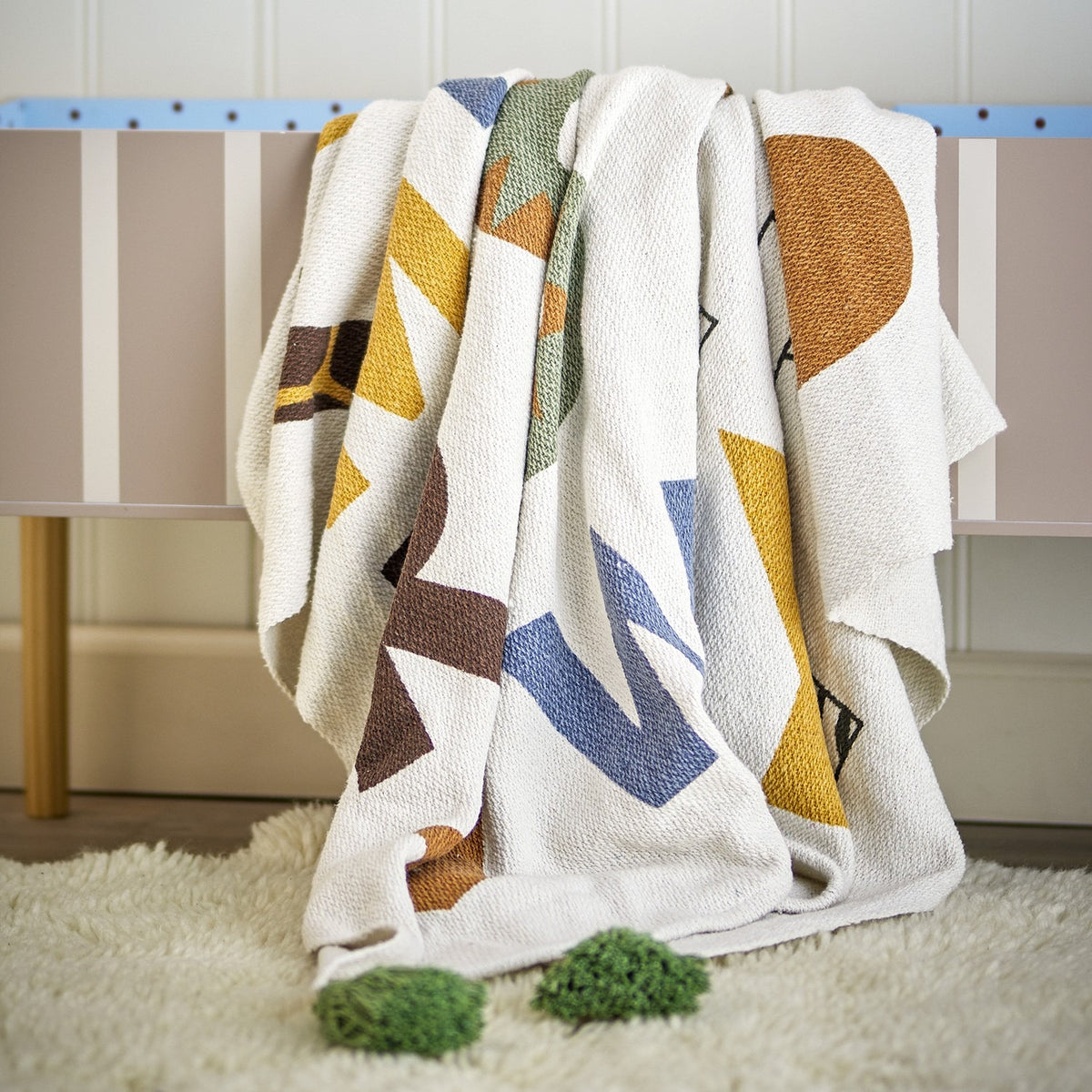 Memmo Cotton Throw in Multi, a cotton blanket with abstract geometric shapes in mustard, blue, green and brown on a cream ground, draped over a striped bench.