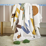 Memmo Cotton Throw in Multi, a cotton blanket with abstract geometric shapes in mustard, blue, green and brown on a cream ground, draped over a striped bench.