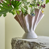 Plier Stoneware Vase 29cm in pink, fan-shaped fluted rim on a flared foot, shown holding green foliage and wildflowers on a stone surface.