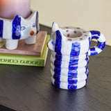 Caty Striped Stoneware Candle Holder in blue, cat-shaped with pointed ears, curled tail handle and ribbed stacked body, shown on a dark table beside a smaller matching holder and books.