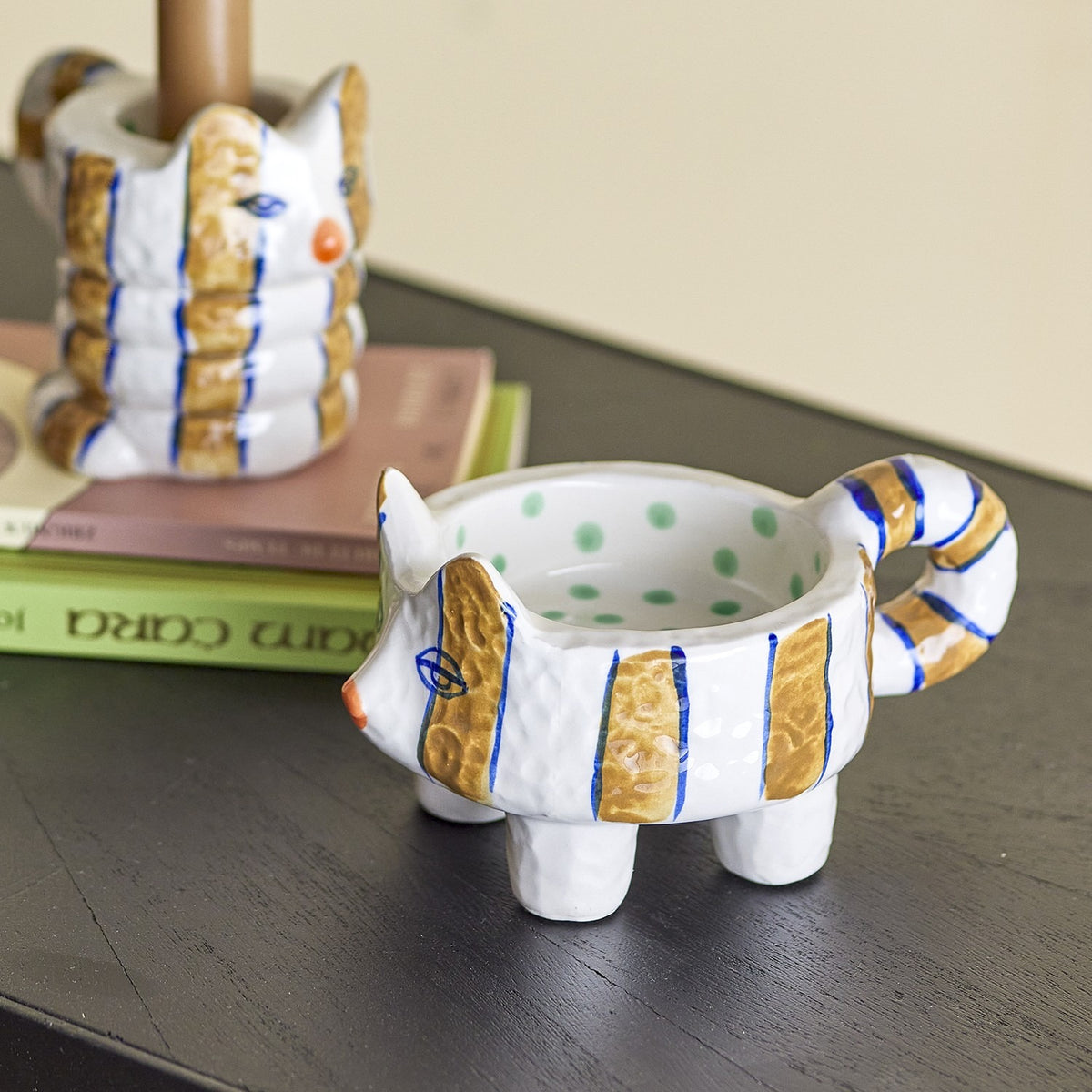 Caty Striped Stoneware Candle Holders Set of 2: cat-shaped stoneware pieces in white with gold and blue stripes, one four-legged with a loop-tail handle, shown on a dark surface beside books.