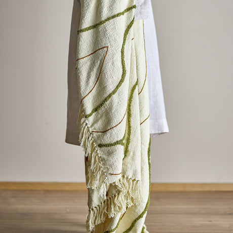 Osasio Cotton Throw in cream draped over a chair, woven cotton with tufted green and stitched rust lines and knotted fringe, shown against a plain light background.