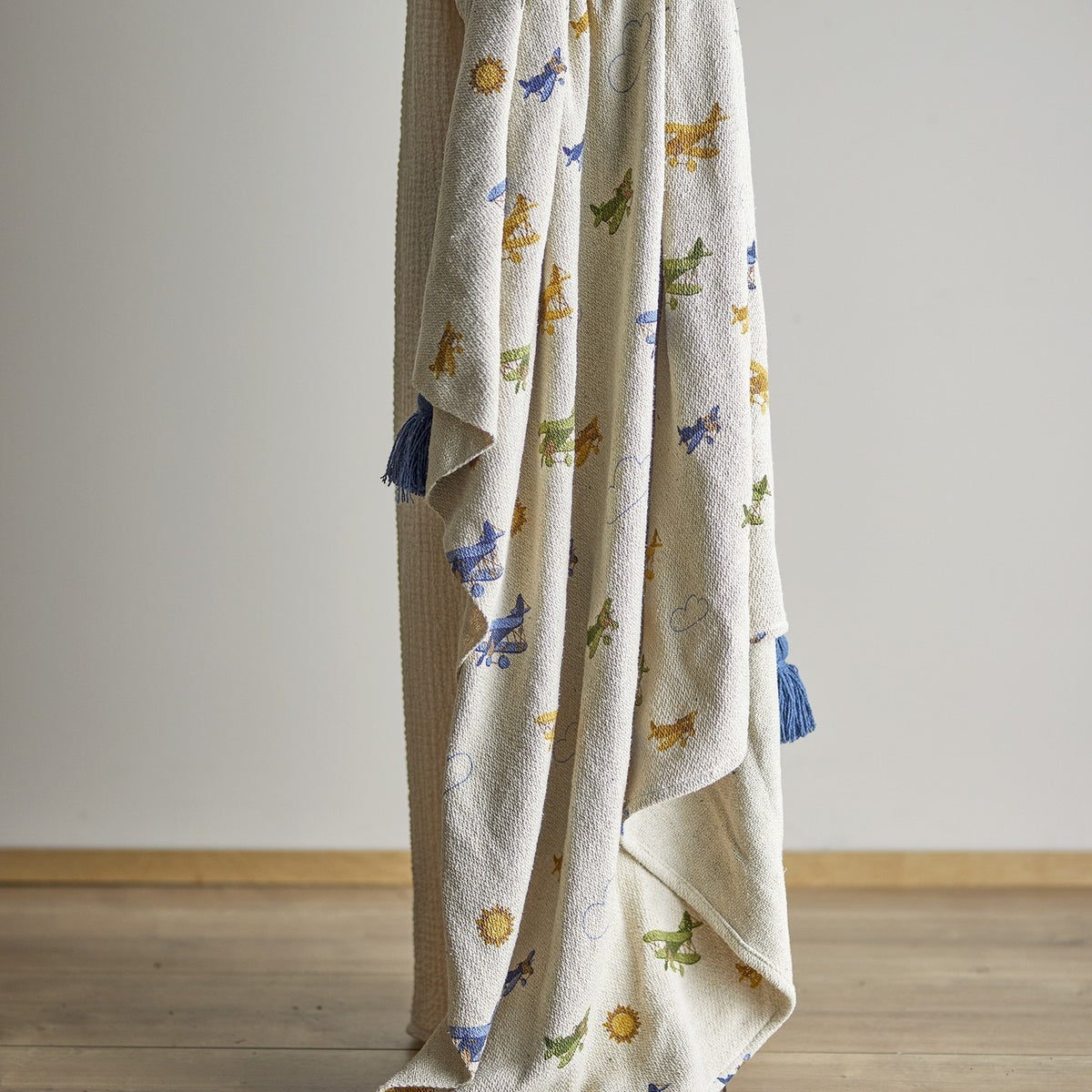 Plane Cotton Throw in beige, draped to show the all-over biplane, cloud, and sun print with chunky blue tassels at the corners, against a plain wall.