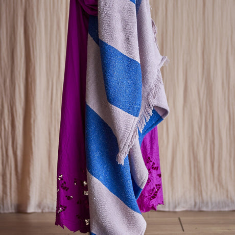 Cadeo cotton throw in blue with wide diagonal pale stripes, fringed edge, draped alongside a magenta fabric against a neutral curtain backdrop.
