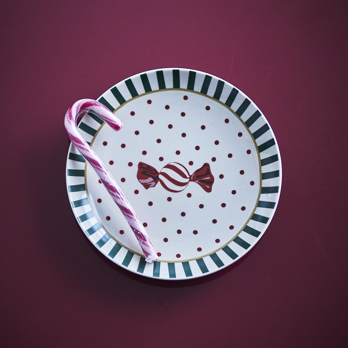Yule Striped Stoneware Christmas Plate in white with green striped rim, red polka dots and a wrapped sweet motif, shown on a maroon background with a candy cane.