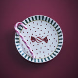 Yule Striped Stoneware Christmas Plate in white with green striped rim, red polka dots and a wrapped sweet motif, shown on a maroon background with a candy cane.
