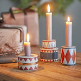 Bitja Geometric Christmas Candle Holders, set of three stoneware pieces in multicoloured zigzag, triangle, and diamond patterns, shown lit with taper candles on a wooden table.