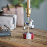 White resin carousel horse candle holder on a red and white striped base with a gold bell-shaped candle cup, displayed on a wooden table with gifts nearby.