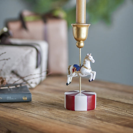 White resin carousel horse candle holder on a red and white striped base with a gold bell-shaped candle cup, displayed on a wooden table with gifts nearby.