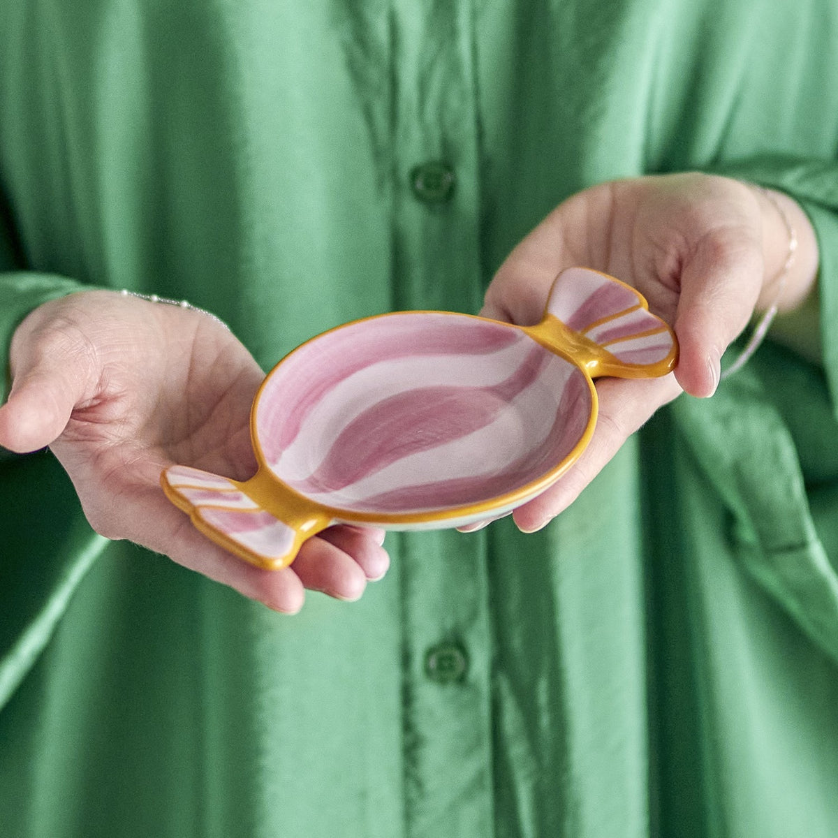Caramella Wrapped Sweet stoneware platter in pink, shaped like a twisted candy wrapper with hand-painted swirls, shown held in two hands against a green shirt.