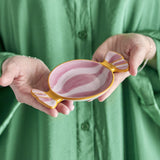 Caramella Wrapped Sweet stoneware platter in pink, shaped like a twisted candy wrapper with hand-painted swirls, shown held in two hands against a green shirt.