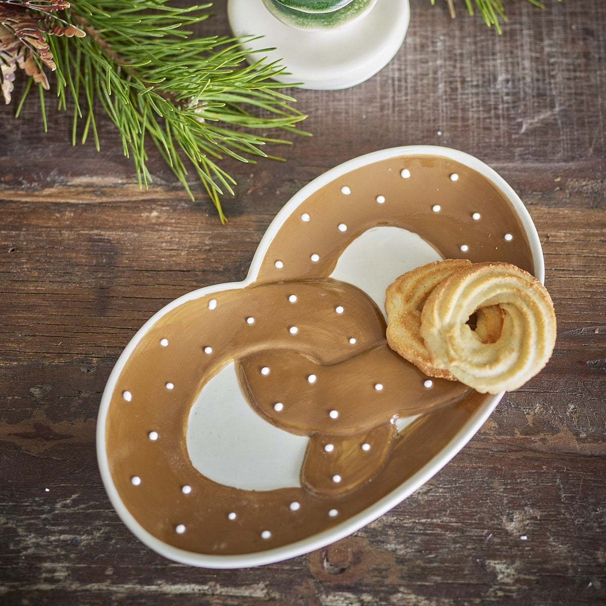 Treats Pretzel Stoneware Christmas Platter in Brown, a glossy pretzel-shaped serving dish with white dot detailing, shown with a pastry on a dark wood table beside pine branches.