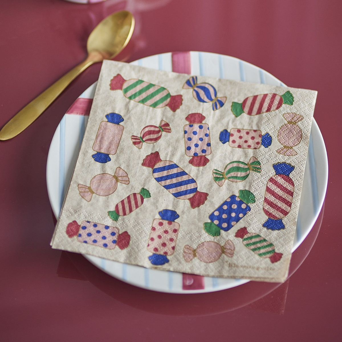 Caramella paper Christmas napkin with multicoloured wrapped-sweet print, folded on a white plate beside a gold spoon on a dark red table.