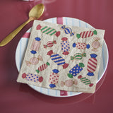 Caramella paper Christmas napkin with multicoloured wrapped-sweet print, folded on a white plate beside a gold spoon on a dark red table.