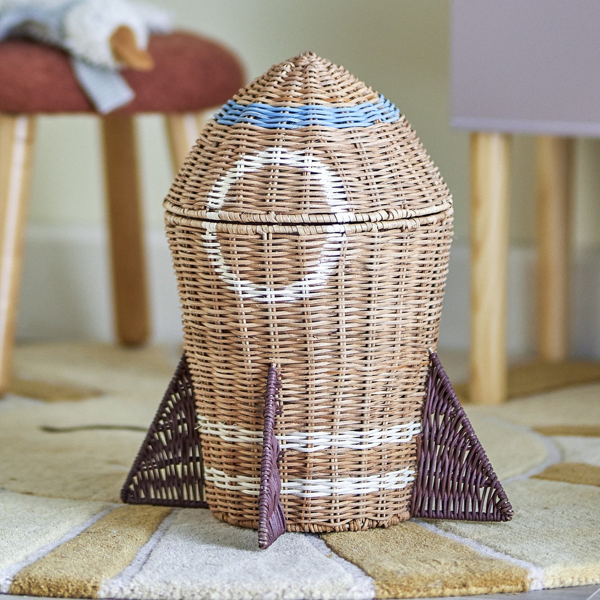Space Rocket Rattan Basket with Lid in natural woven finish, featuring a pointed lid, white porthole detail, blue band and triangular base fins, shown on a rug in a nursery setting.