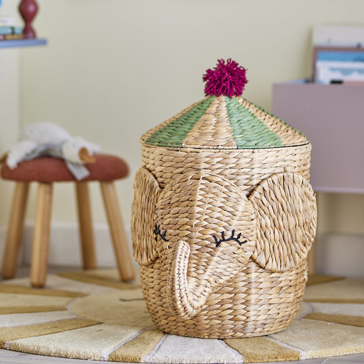 Bobo elephant water hyacinth storage basket with lid, natural wicker weave with curled trunk, stitched eyelashes, and green-striped conical lid topped with a pink pompom, shown in a nursery setting.