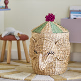 Bobo elephant water hyacinth storage basket with lid, natural wicker weave with curled trunk, stitched eyelashes, and green-striped conical lid topped with a pink pompom, shown in a nursery setting.
