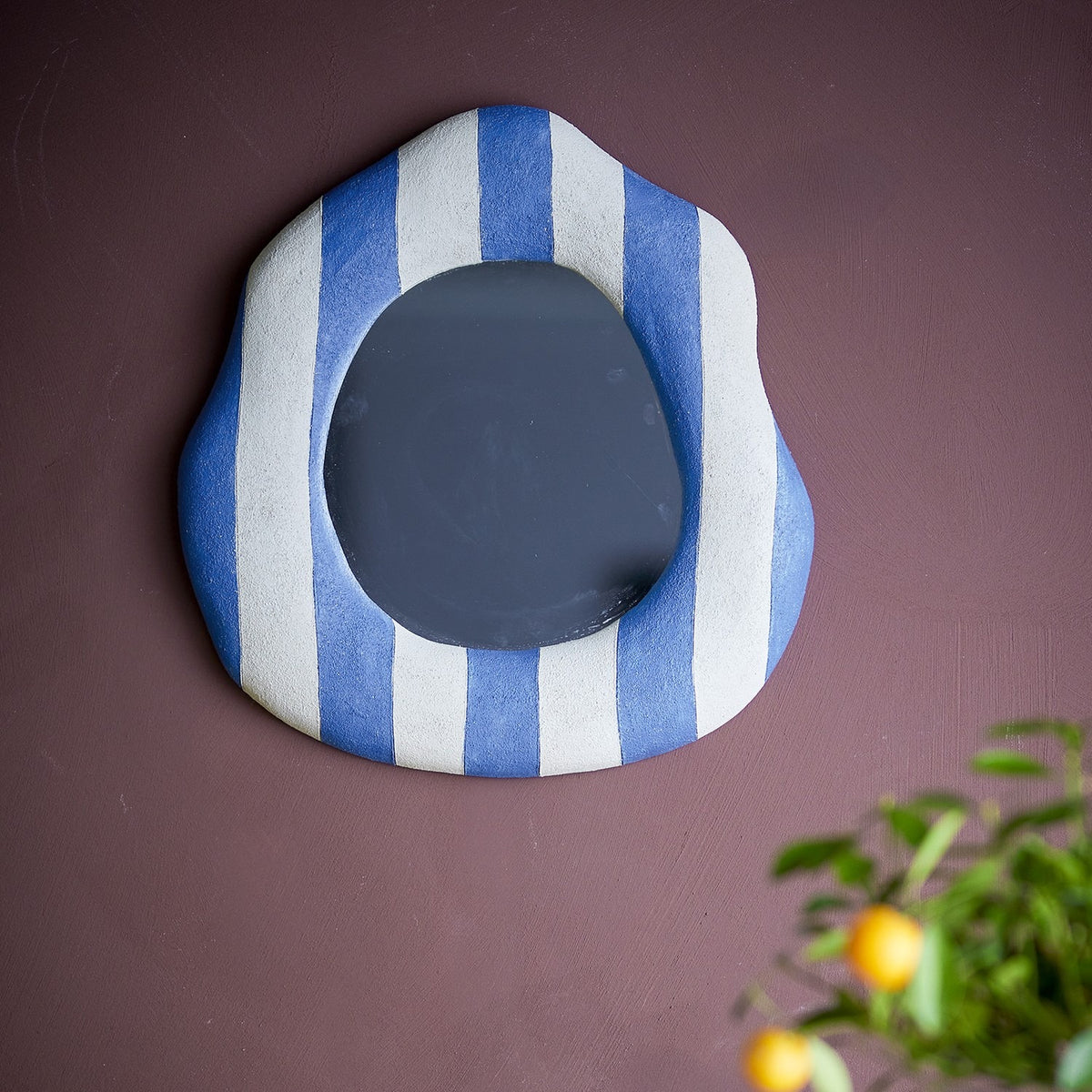 Travis Striped Resin Wall Mirror 39x38cm in Blue, an irregular rounded frame with broad blue and white stripes, mounted on a dark wall near a potted plant.