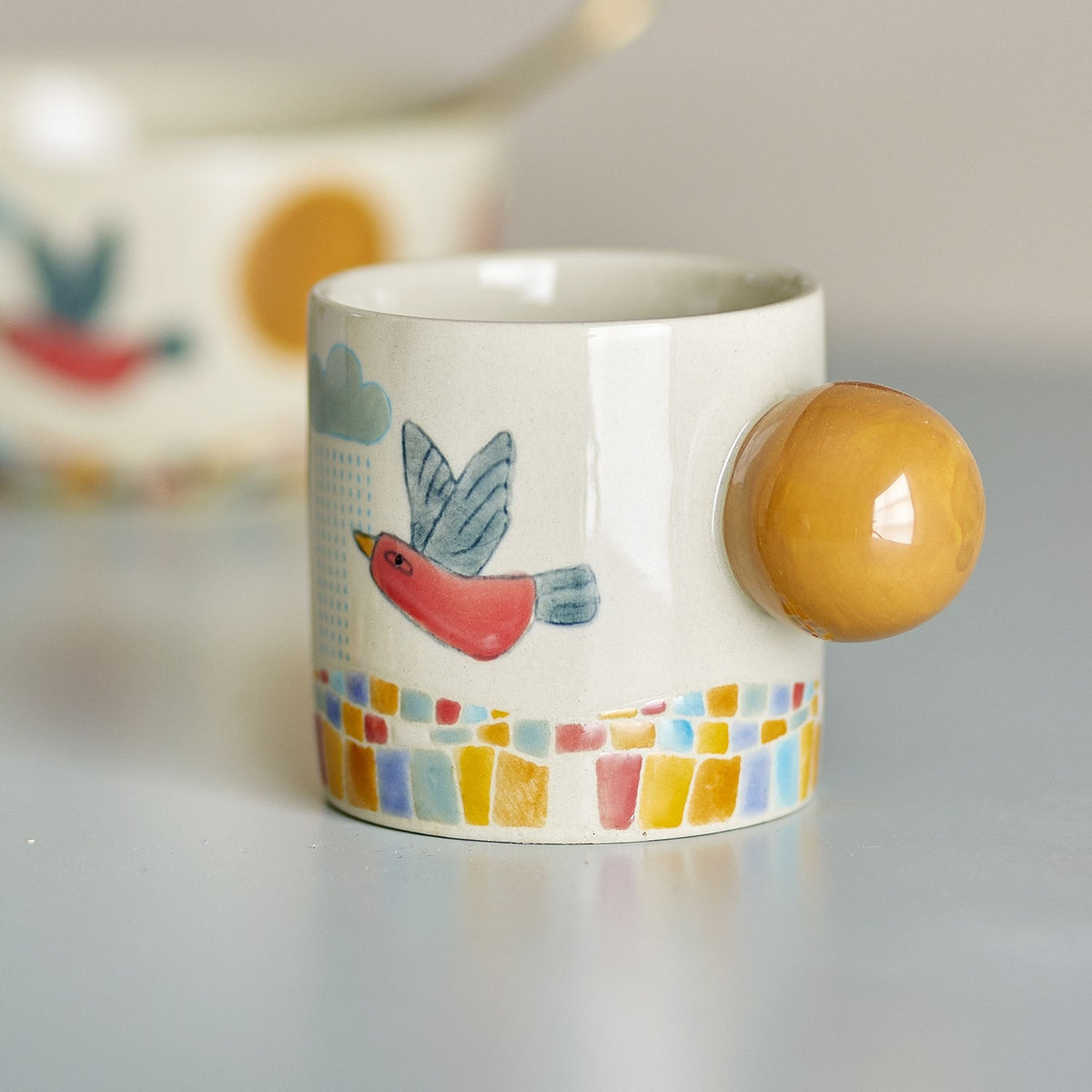 Colorine Stoneware Cup 155ml in multi, featuring a hand-painted bird under a rain cloud, mosaic-pattern base, and a round mustard-yellow ball handle, shown on a light grey surface.