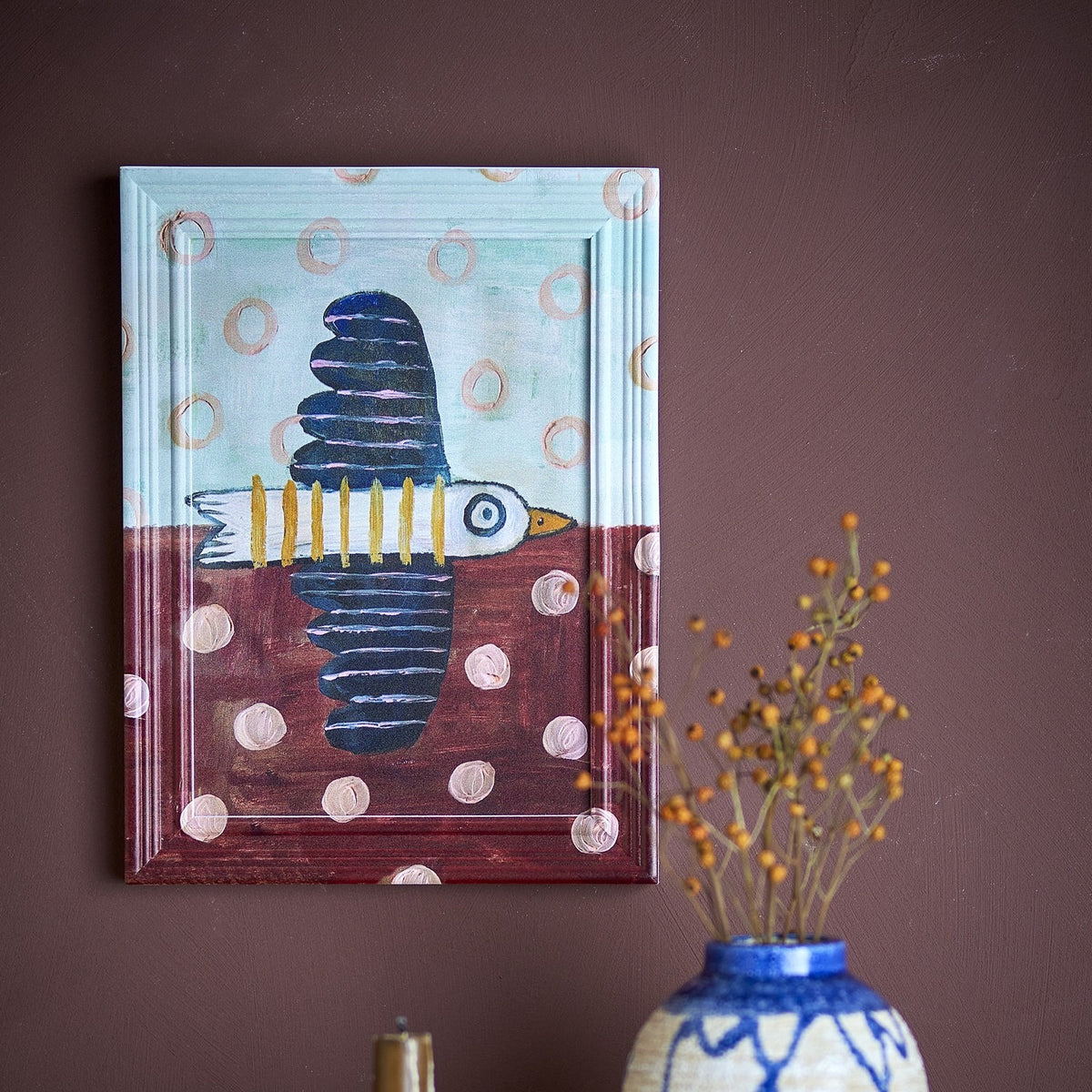 Brett birdy Wooden Framed Print in multicoloured, showing a bird in flight with scalloped wings, rings and dots, in a moulded wood frame on a dark wall above a vase.