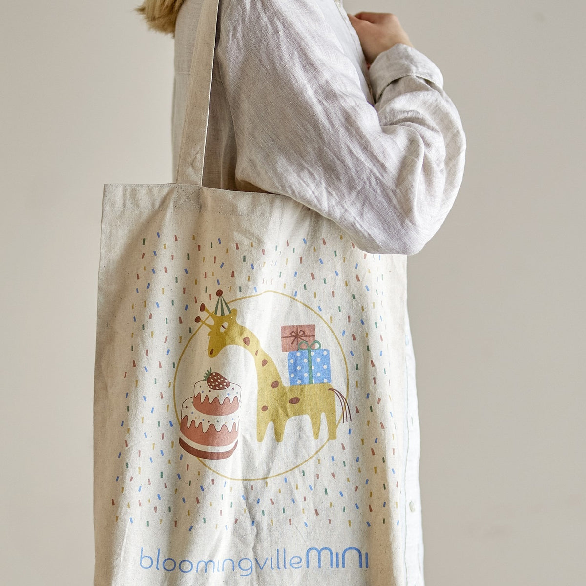 Celebrate Cotton Tote Bag in beige, shown carried over the shoulder, printed with a giraffe in a party hat beside a cake and gifts, surrounded by confetti flecks.